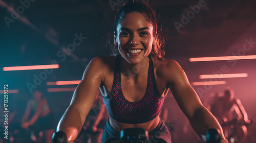 A determined-looking woman on a cycling bike in a spin class, catching the camera with a quick, powerful smile.
