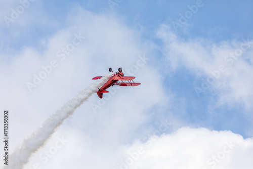 A restored World War II era red biplane airplane performing aerobatic maneuvers in the sky during an air show event, leaving a trail of smoke against a cloudy background in Hillsboro, Oregon, USA