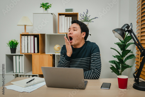sleepy man yawning while working with laptop computer at home office