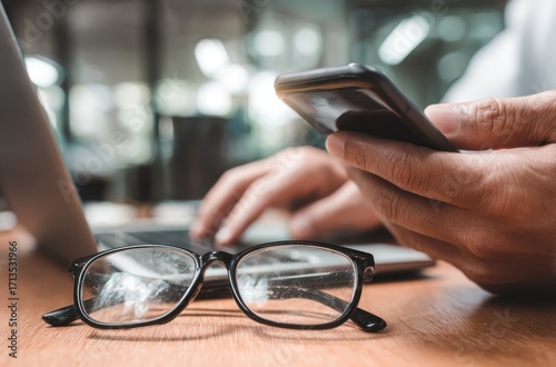 Person using a phone, with glasses, and a laptop on a wooden table. Natural light creates a warm and focused setting for productivity