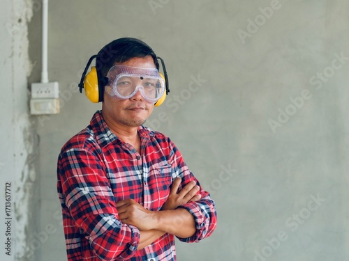 Asian carpenter wearing a red plaid shirt, standing with arms crossed looking at the camera, wearing clear glasses and earmuffs, personal protective equipment, work safety