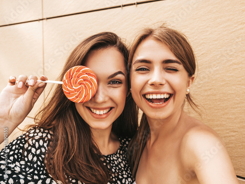 Two young smiling hipster women in summer clothes. Female taking selfie self portrait photos on smartphone.Models posing on street background near yellow wall.Eating candy lollipop