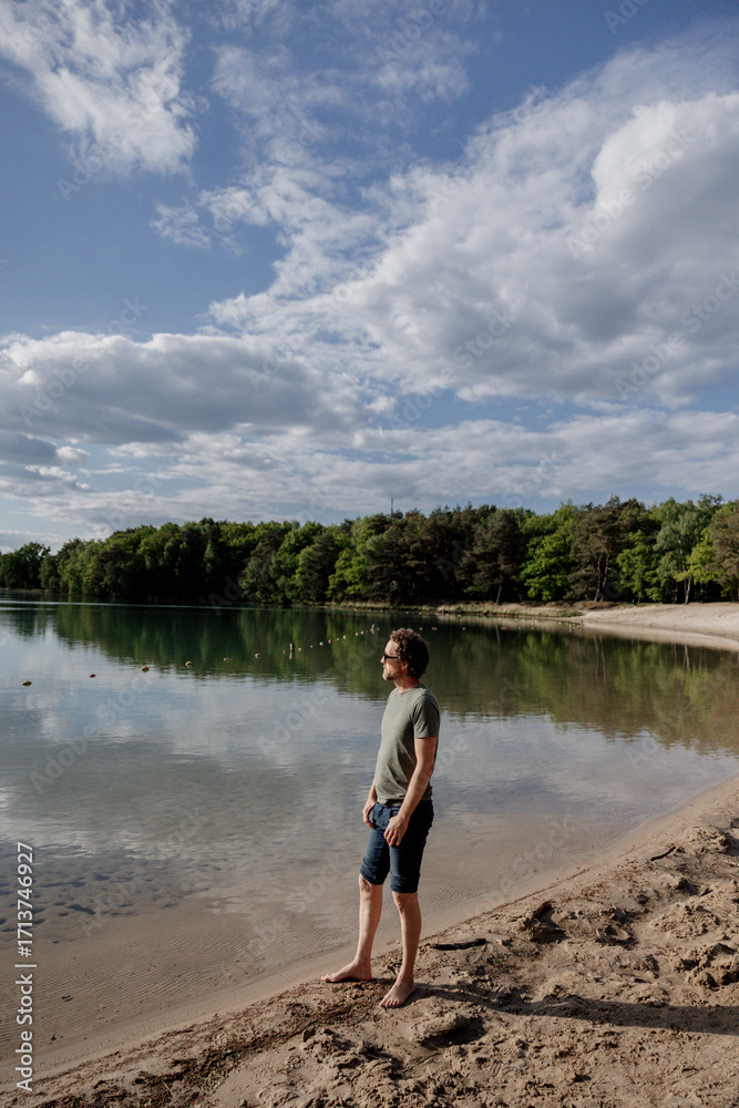 Fototapeta premium Person daydreaming by a calm lake on sandy beach.