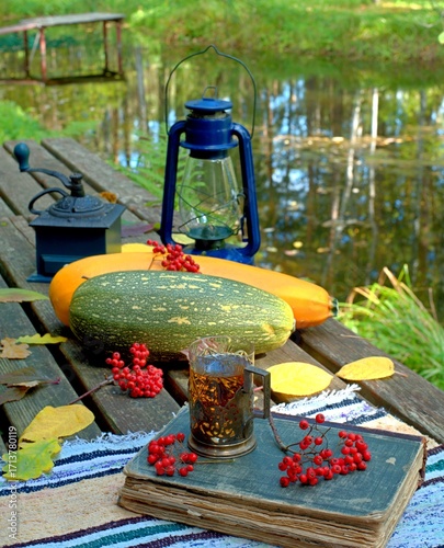 Autumn composition with zucchini, glass of tea, old book and kerosene lamp. Autumn retro home decor with lake in the background.