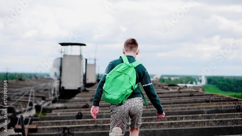 A boy walks alone on the roof of a tall house. He walks on concrete among the wires