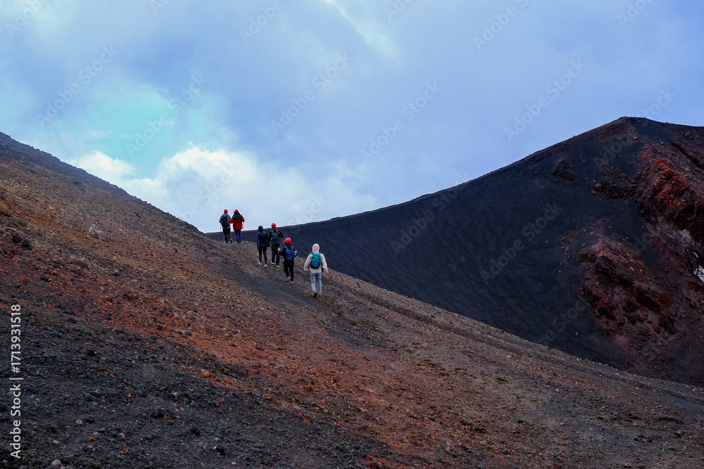 Obraz premium Group of hikers exploring volcanic slopes with dramatic red and black ash textures under cloudy sky, adventure trekking on active mountain terrain, perfect for travel, geology, and outdoor exploration