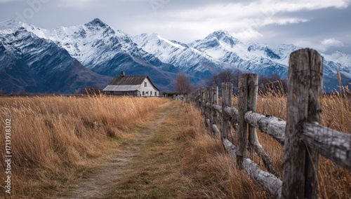 Rustic farmhouse nestled in a valley, beside a path lined with a weathered wooden fence, facing snow-capped mountains