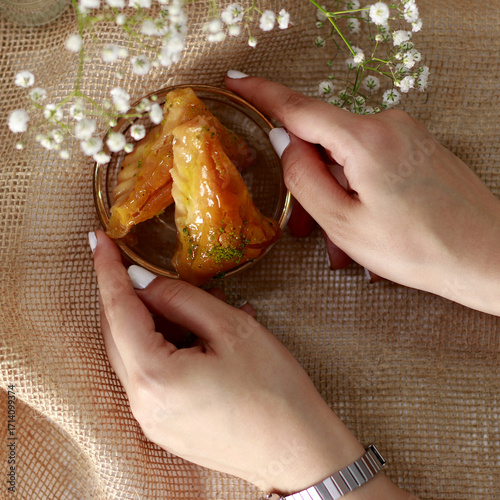 Obraz na plátne Hands holding small plate with slice of golden syrupy pastry placed on rustic fabric with flowers