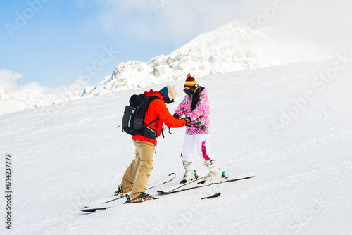 Male Ski instructor in red jacket assisting hold hands beginner female adult skier in pink outfit on snowy mountain slope, concept of ski school, training, winter tourism and lifestyle