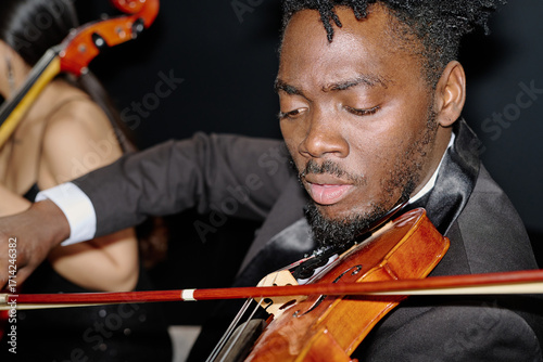 Black young adult man playing violin in formal attire during orchestral performance, focusing intently on instrument, with  woman partially visible in background