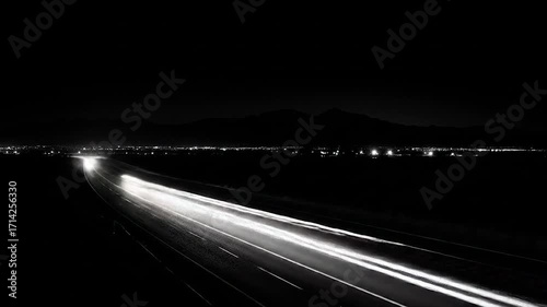 Night highway, illuminated by streaks of car lights, stretches across a landscape under dark mountains.