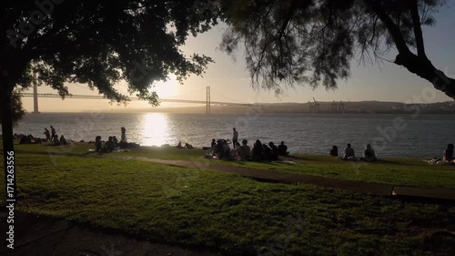 Lisbon Portugal. People relaxing on the riverside lawn with the 25th of April Bridge, sailboats floating on the Tagus River at sunset. Park by the riverside