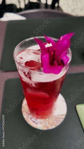 Close-up of a refreshing red drink with ice, served in a tall glass and decorated with a vibrant pink flower. Bright summer beverage with floral garnish on a restaurant table