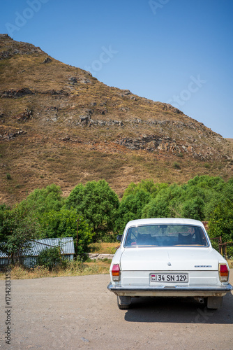 Classic white car parked on a rural road with rocky mountains and green trees in the background. A nostalgic travel scene blending vintage automotive charm with natural countryside