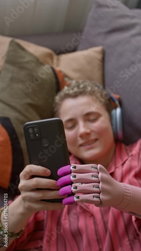 Vertical top view shot of smiling young woman looking at smartphone screen while lying on bed at home with focus on prosthetic hand holding phone