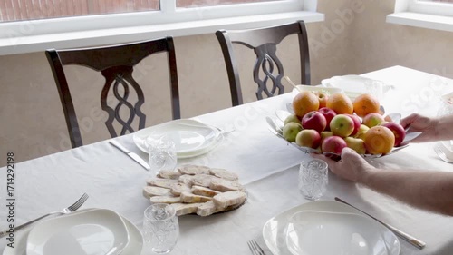 Family dinner table setting with white tablecloth and plates. Man placing a large bowl of apples and oranges on a wooden table.