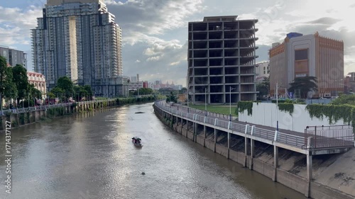 Riverside view in Phnom Penh, Cambodia, with mix of modern high-rise towers, and unfinished concrete building. Small boat moves along, highlighting the blend of urban development and traditional life