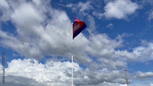 Cambodian national flag flying on a tall pole in Phnom Penh. The flag waves gracefully against a vivid blue sky with dramatic white clouds and birds in flight, symbolizing pride, identity, and freedom