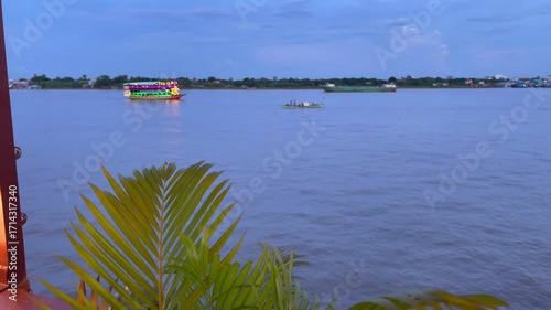 Evening view of the river in Phnom Penh, Cambodia, showing a brightly decorated tourist boat with colorful neon lights alongside a smaller canoe carrying locals