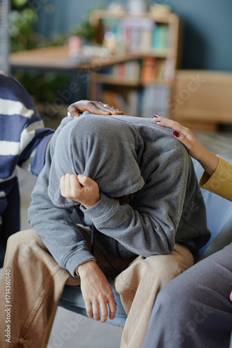 Teenager boy with hood covering head sitting with face in hands, receiving comfort from multiethnic group with supportive hands on shoulder and back, emotional support concept