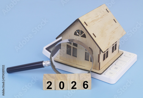 Wooden house model on calculator and cubes with numbers 2026 under magnifying glass. Real estate price in year 2026.