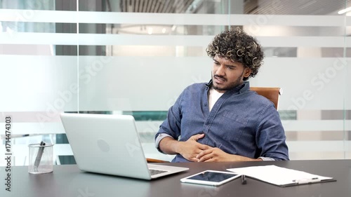 Unhealthy young businessman feels stomach pain while working on a laptop sitting at workplace in business office. Sick worker has heartburn, gastritis or poisoning, suffers from spasms or constipation