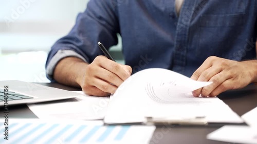 Close up of a male's hand signing documents at a desk at a workplace in a modern business office. The boss or manager looks through the folder with documentation and signs the contract with a pen