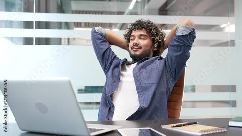 Happy businessman finished work on laptop while sitting at desk at workplace in a modern business office. Smiling satisfied man puts his hands behind his head, stretches himself in a chair. Work done