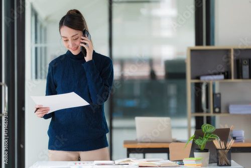 Image of an Asian businesswoman holding a document report and using a cellphone while working in an office.