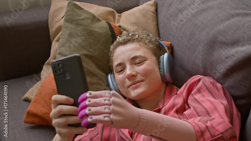 Top view shot of smiling young woman with prosthetic hand holding smartphone above while watching videos relaxing on couch