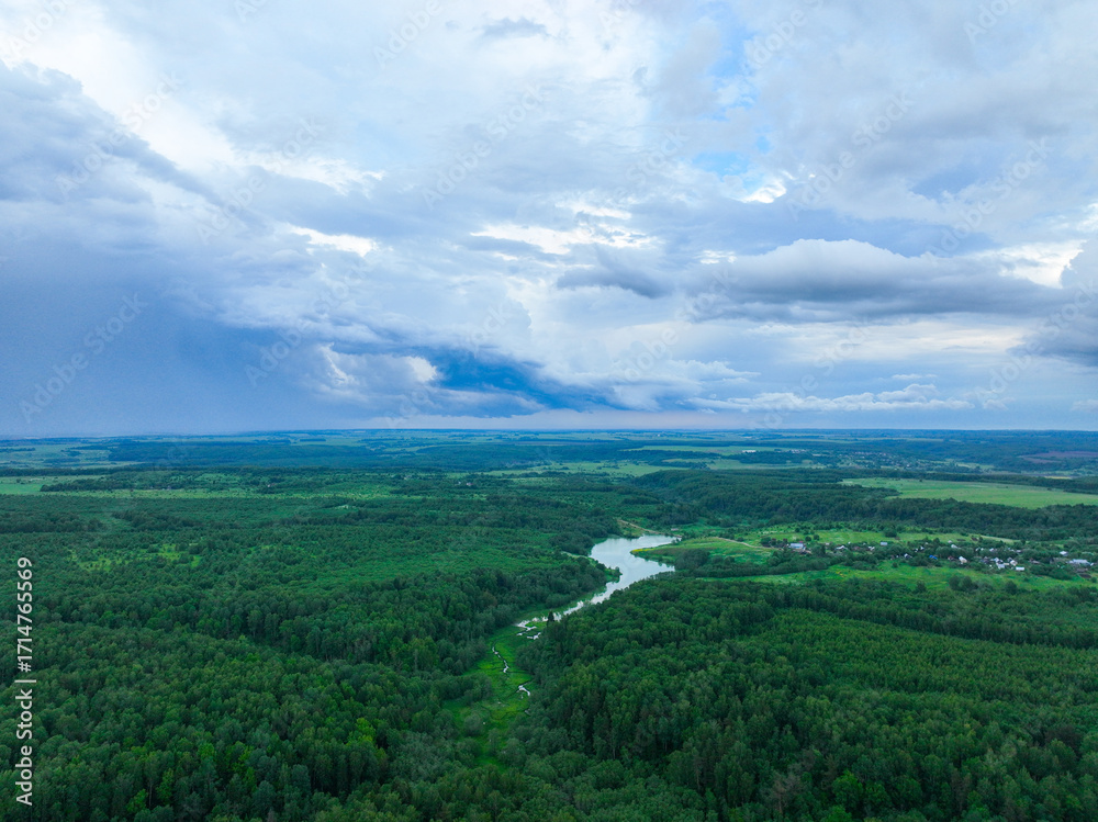 Obraz premium A wide aerial view of a winding river in the middle of a dense green forest, with a stormy, dark cloudscape above it.