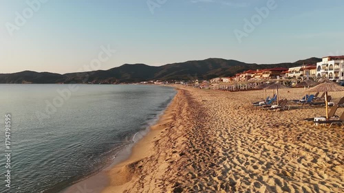 High-resolution aerial drone image showcasing a picturesque beachfront resort scene. Neatly arranged white umbrellas and lounge chairs line the golden sandy shore, while colorful hotel buildings with 