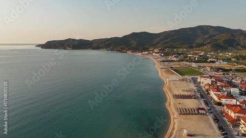 High-resolution aerial drone image showcasing a picturesque beachfront resort scene. Neatly arranged white umbrellas and lounge chairs line the golden sandy shore, while colorful hotel buildings with 