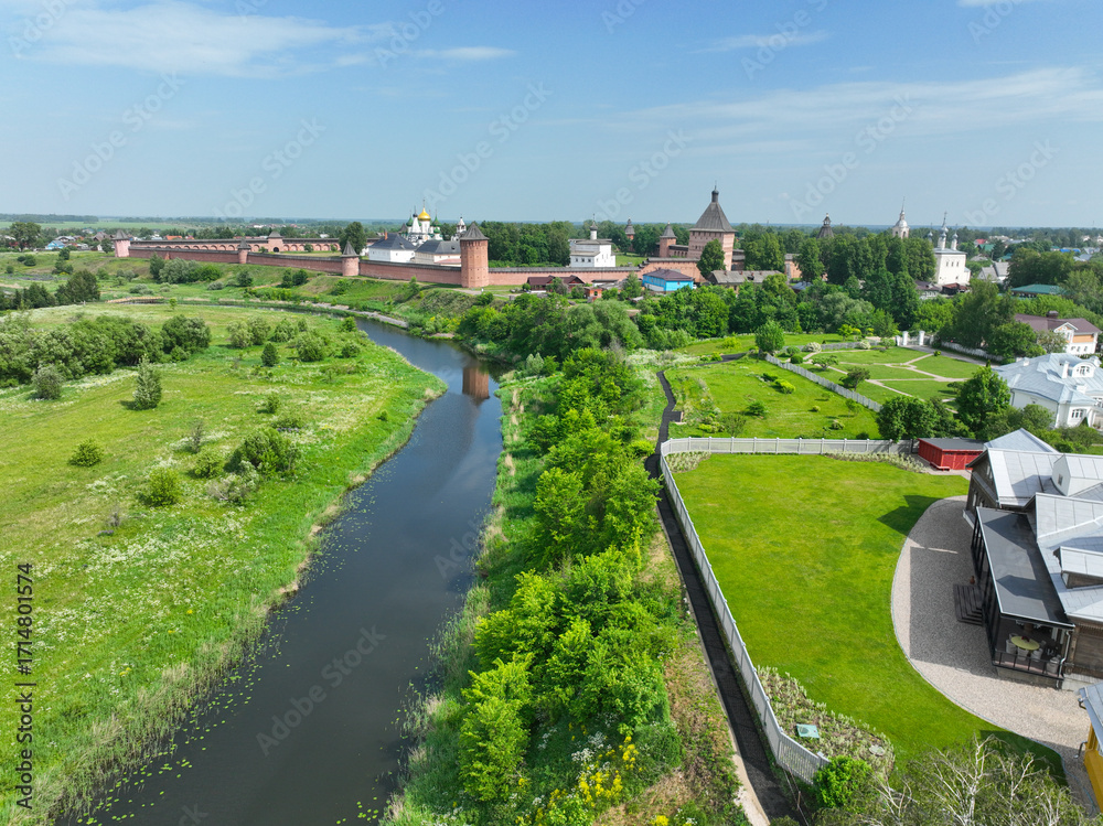 Fototapeta premium An aerial view of a large monastery complex with red brick walls and towers, next to a small river and green fields.