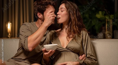 Caring Husband Affectionately Feeds His Smiling Pregnant Wife from a Bowl, Sharing a Tender Moment on the Couch, Anticipating Their Future Family