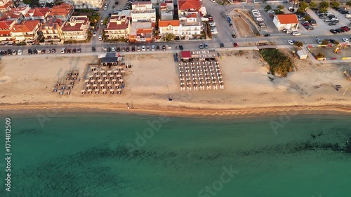 High-resolution aerial drone image showcasing a picturesque beachfront resort scene. Neatly arranged white umbrellas and lounge chairs line the golden sandy shore, while colorful hotel buildings with 