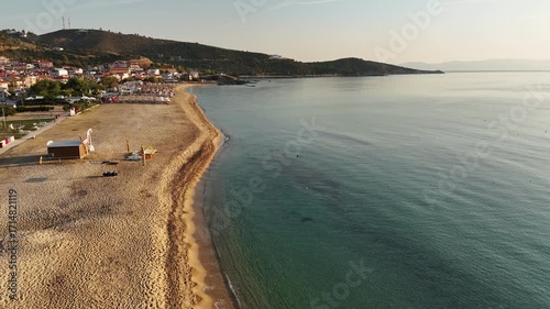 High-resolution aerial drone image showcasing a picturesque beachfront resort scene. Neatly arranged white umbrellas and lounge chairs line the golden sandy shore, while colorful hotel buildings with 