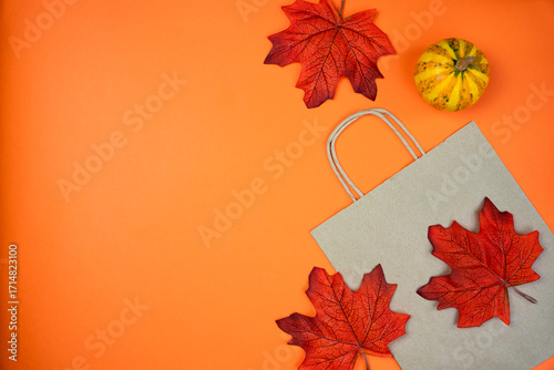Autumn flatlay composition with paper shopping bag, autumn maple leaves and small pumpkin on an orange background. Fall shopping. Thanksgiving sales concept. Top view. composition. Copy space.