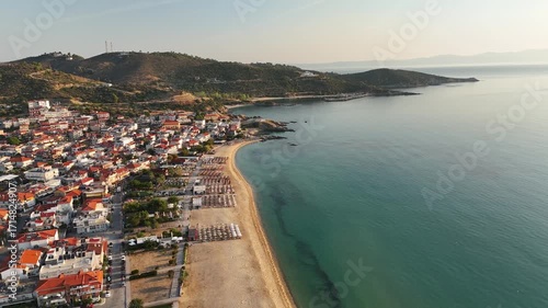 High-resolution aerial drone image showcasing a picturesque beachfront resort scene. Neatly arranged white umbrellas and lounge chairs line the golden sandy shore, while colorful hotel buildings with 