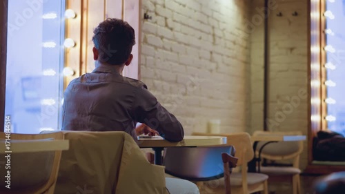 young man sitting alone at round table near window in empty cafe, coat draped over chair, back view, soft lights glowing, waiting in quiet contemplative mood amid minimal brick interior