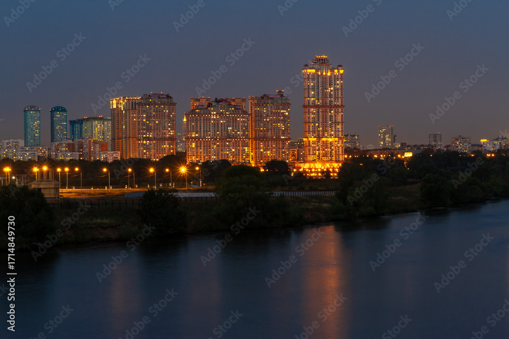 Obraz premium Night view of Moscow River from high bank in Strogino, with modern residential buildings of Shchukino reflected in dark water. Peaceful urban landscape. Moscow, Russia.