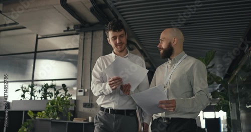 Two male coworkers walk together in a modern office holding documents and discussing work projects teamwork collaboration professional atmosphere