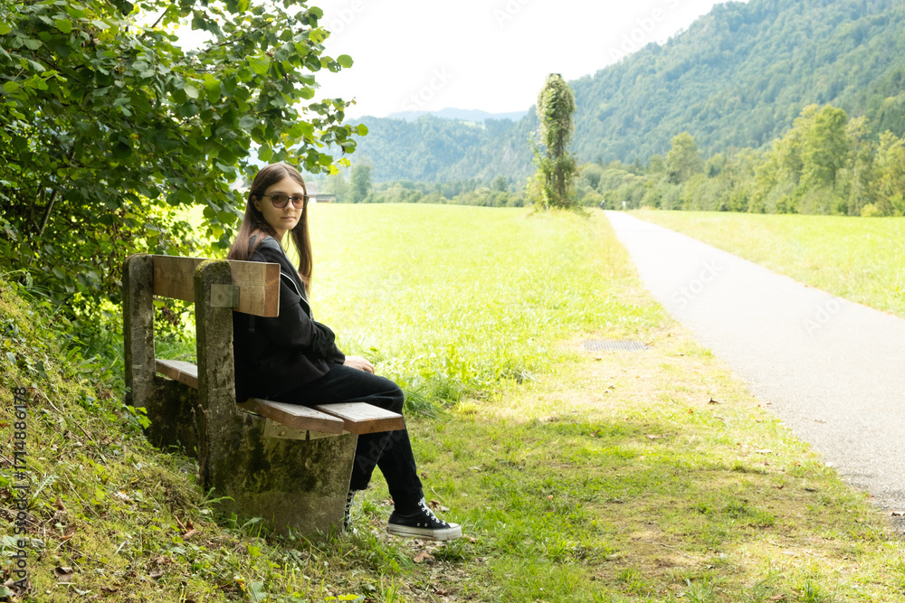 Fototapeta premium Teenage girl sitting on a bench in nature, Alps and meadows of Europe