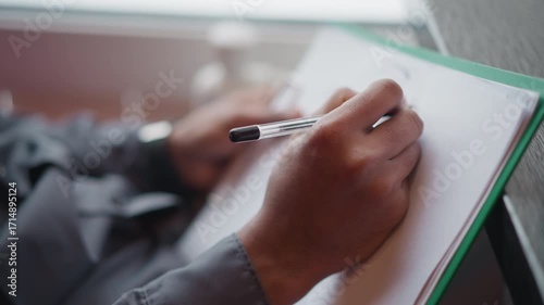 Immigrant writing with pen on white paper clipped to green folder near window, close-up of hands and focus on movement of pen, suggesting concentration, creativity, or preparation in calm indoor