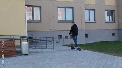 A young man rides an electric scooter along a paved path in an urban park with modern apartment buildings in the background.