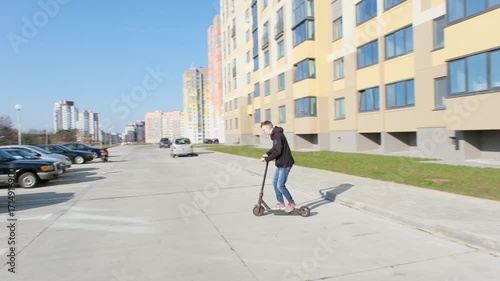 A young man rides an electric scooter along a paved path in an urban park with modern apartment buildings in the background.