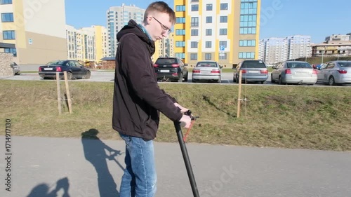 A young man rides an electric scooter along a paved path in an urban park with modern apartment buildings in the background.