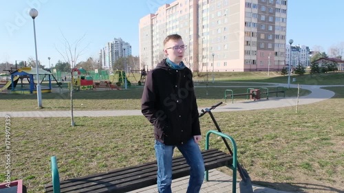 A young man rides an electric scooter along a paved path in an urban park with modern apartment buildings in the background.