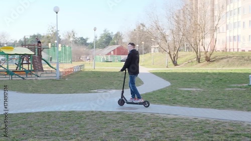 A young man rides an electric scooter along a paved path in an urban park with modern apartment buildings in the background.