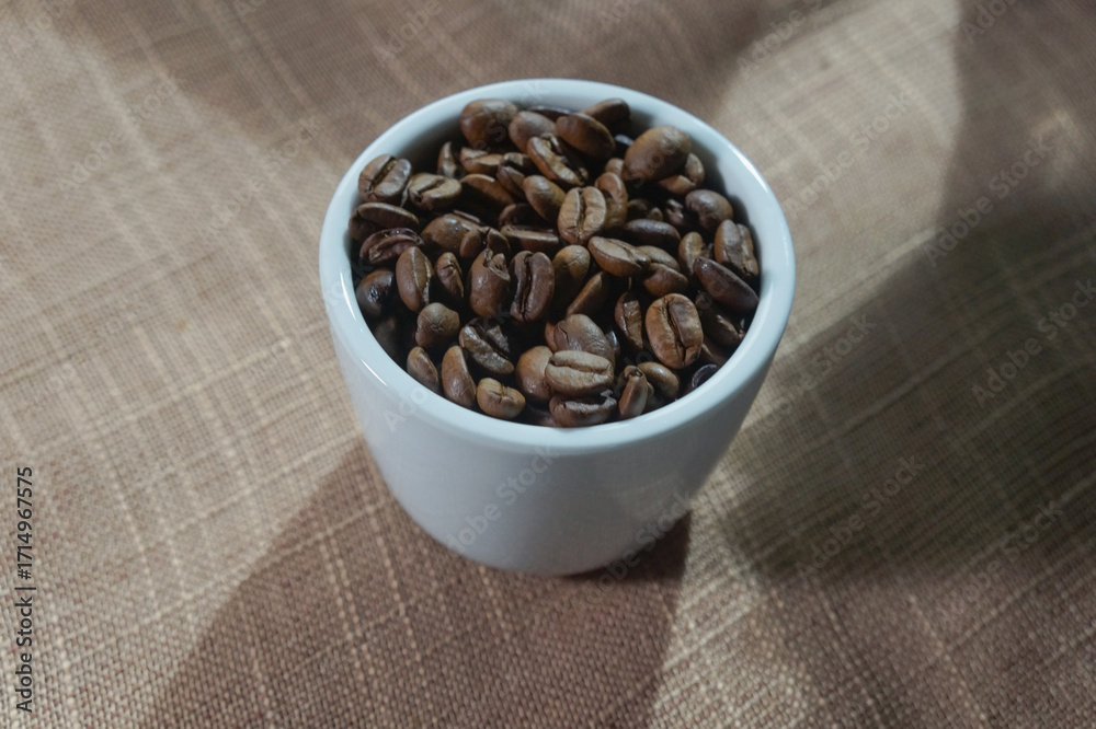 Fototapeta premium White ceramic mug filled with roasted coffee beans on a rustic brown cloth